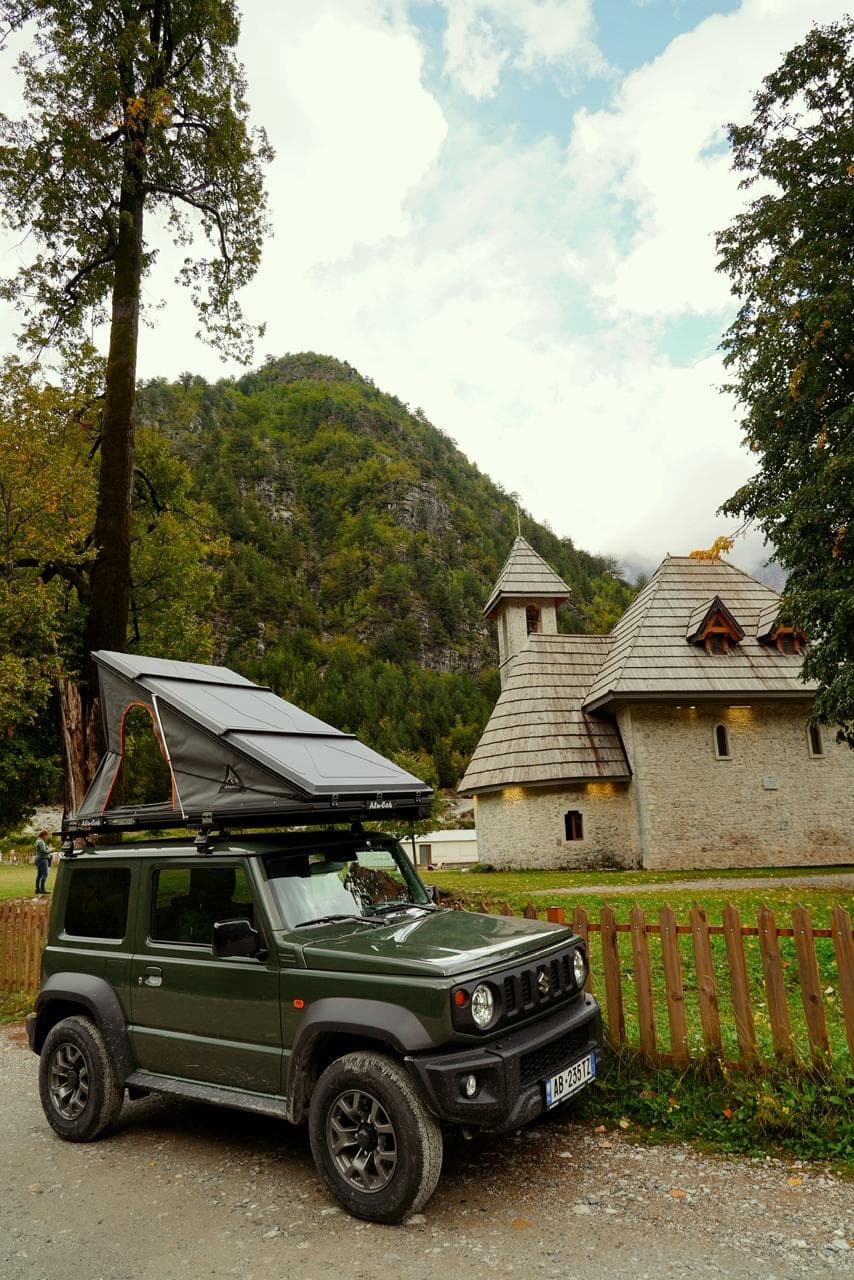 Green Jimny with rooftop tent parked beside a traditional Albanian mountain chapel