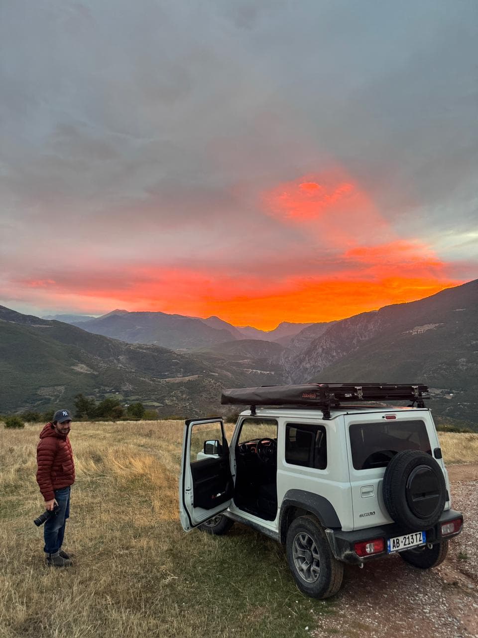 Expert guide beside a Suzuki Jimny under a fiery red Albanian sunset
